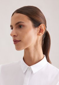 Femme aux cheveux bruns attachés en queue de cheval basse, portant une chemise blanche à col, regardant vers la gauche sur un fond uni clair.