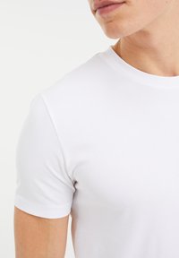 Camiseta blanca de algodón con cuello redondo, mangas cortas y textura suave, que presenta un diseño simple sin patrones ni acentos.