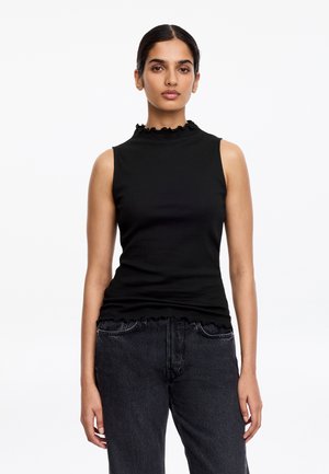 Femme aux cheveux foncés coiffés en chignon bas, portant un haut noir sans manches avec des bords volantés et un jean taille haute foncé, se tenant devant un fond blanc.