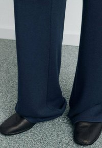 Pantaloni sport largi albastru marin, confecționați dintr-un material moale, asortați cu pantofi slip-on din piele neagră pe un podea texturată gri.