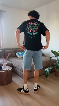 Άτομο με μαύρο γραφικό μπλουζάκι και ανοιχτό γαλάζιο σορτς στέκεται σε ένα σαλόνι με καφέ καναπέ, ξύλινο τραπέζι και πράσινο φυτεμένο φυτό.
