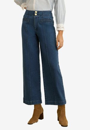 Blauwe jeans met hoge taille en wijde pijpen, met decoratieve gevlochten zakken, gecombineerd met beige suède enkellaarsjes met blokhak en een gestreept bloesje met kanten manchetten.