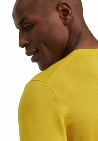 Gelber Strickpullover mit Rundhalsausschnitt, mit nahtlosem Schulterdesign und glatter Textur, seitlich gezeigt vor einem neutralen Hintergrund.