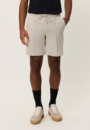 Pantaloni scurți atletici bej, realizați dintr-un material neted, cu talie reglabilă cu șnur, având cusături laterale și un logo subtil. Purtate cu șosete negre și adidași bej.