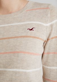 Beige svetr s horizontálními pruhy v bílé, světle růžové a oranžové barvě. Na levé hrudi se nachází malé logo ptáka v barvě maroon. Měkká pletená textura.