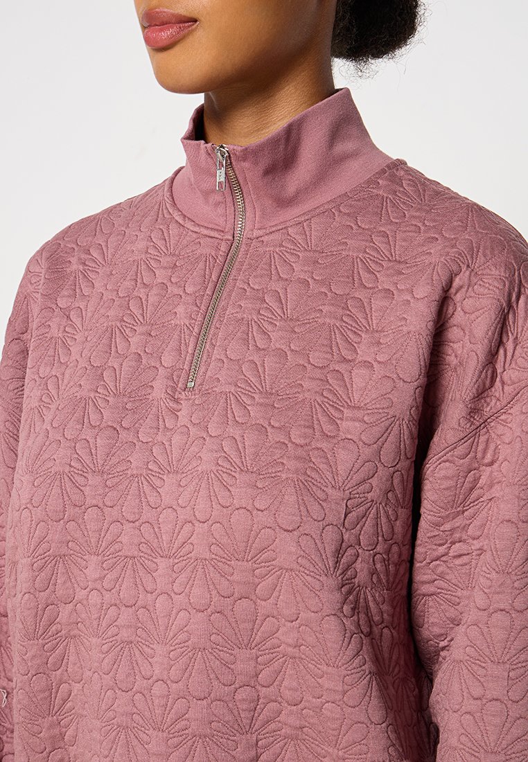 Fleece-Pullover in sanftem Rosa mit hohem Kragen und Halbreißverschluss, ausgestattet mit einem geprägten floralem Muster für zusätzliche Textur und Details.