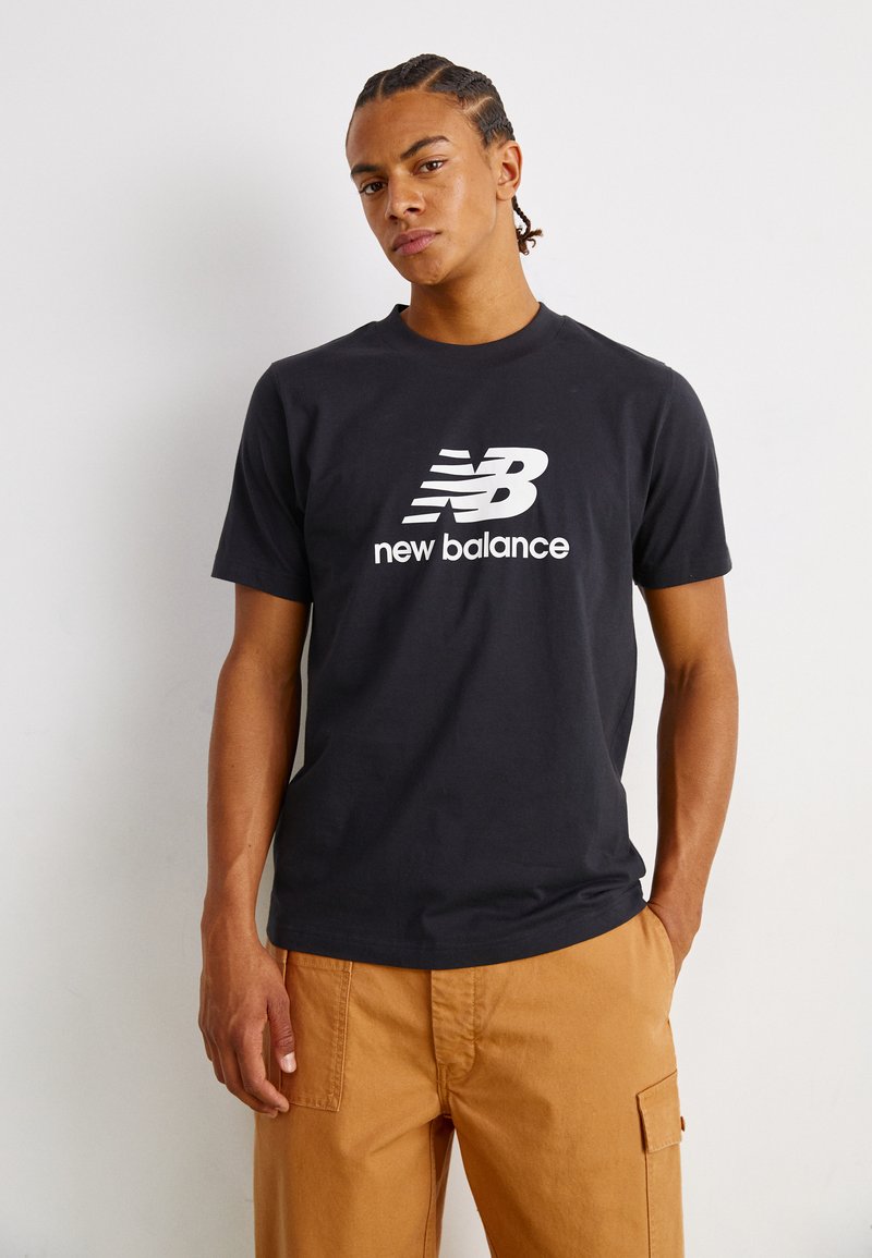 Black cotton t-shirt featuring a white "New Balance" logo. Short sleeves and a round neckline. Paired with tan cargo pants.