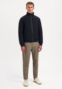 Marineblauwe pufferjack met een opstaande kraag en grijze rits, gecombineerd met khaki broeken en witte sneakers. Soepele textuur, minimalistisch ontwerp.