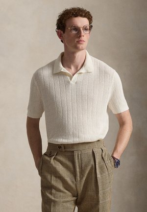 Young man with curly hair wears round glasses, cream knit short-sleeve polo, brown plaid trousers, and a wristwatch, standing with hands in pockets.