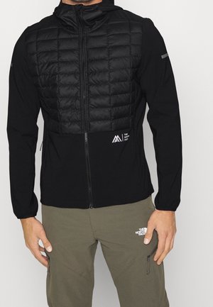 Homme portant une veste matelassée noire zippée à capuche et un pantalon cargo vert olive, tous deux avec les logos The North Face.