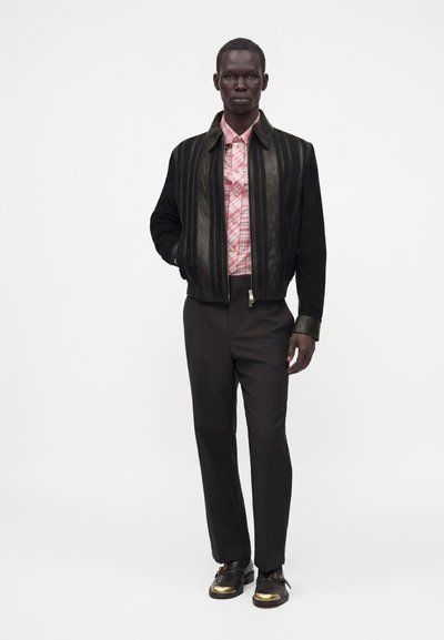 Homme portant une veste bomber noire avec des détails à motifs, une chemise à carreaux rose boutonnée, un pantalon noir et des chaussures noires avec des accents dorés, debout droit.