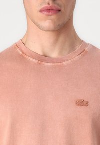 Tricou din bumbac de culoare roz deschis, cu guler rotund, având un mic logo brodat texturat pe partea stângă a pieptului. Finisaj simplu și neted al materialului.