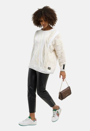 Frau mit lockigem Haar, die einen weißen strukturierten Pullover, schwarze Lederhose, weiße Sneakers trägt und eine braune gemusterte Handtasche an einer Goldkette hält.