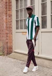 Groen-wit gestreepte polo shirt, bordeauxrode broek met witte en rode zijkanten, witte sneakers en een lichtroze pet. Staande tegen een bakstenen muur.