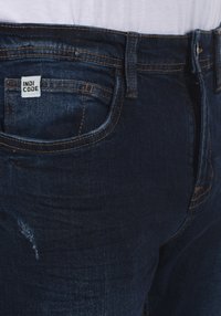 Mørkeblå denimjeans med let slidte detaljer, med et mærketikette på frontlommen og kontraststing accenter.