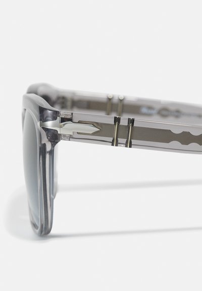 Óculos de sol cinzentos e transparentes com uma armação robusta, apresentando detalhes metálicos prateados e um acabamento liso e brilhante. As lentes são escuras e reflexivas.