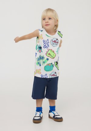 Camicetta bianca senza maniche con stampe grafiche colorate; pantaloni shorts blu; sneakers scure con accenti bianchi; calzini blu.