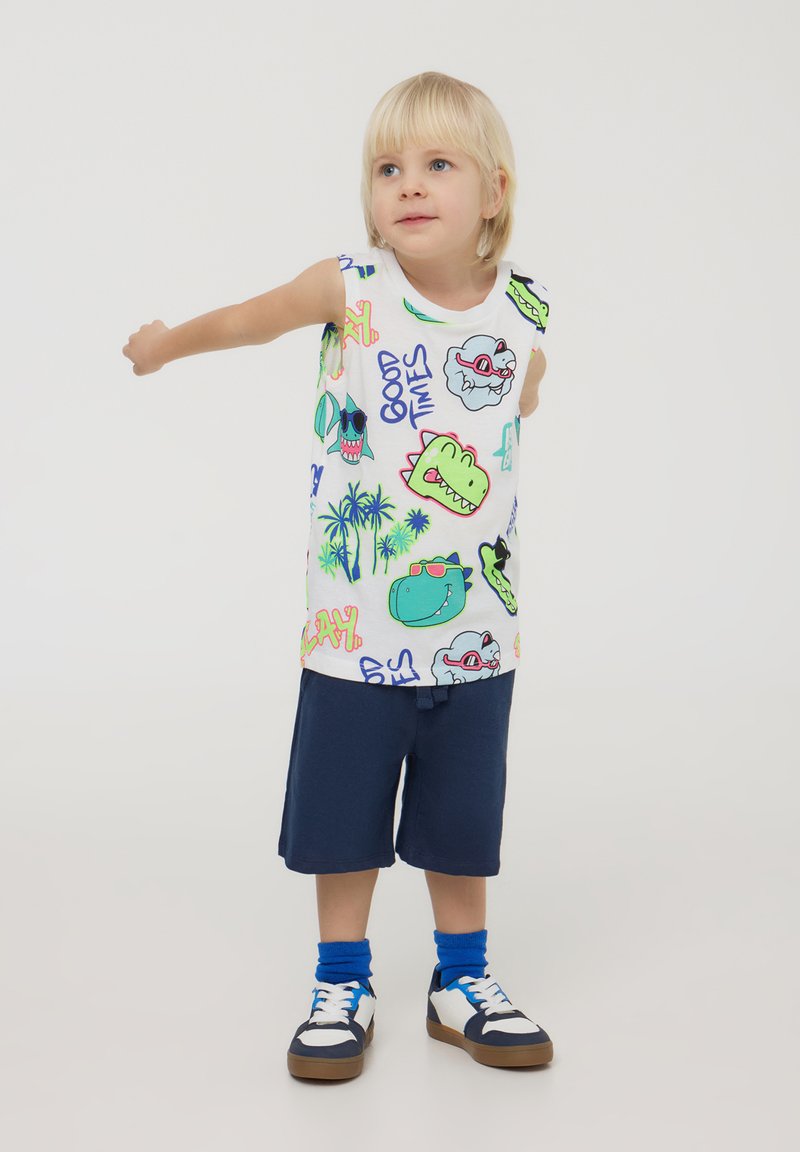 Camicetta bianca senza maniche con stampe grafiche colorate; pantaloni shorts blu; sneakers scure con accenti bianchi; calzini blu.