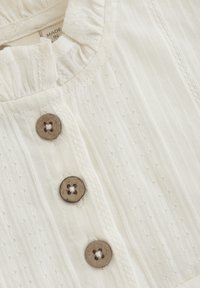 Weißes strukturiertes Stoffoberteil mit rüschigem Ausschnitt und drei braunen Holzknöpfen mit floralen Ausschnitten. Dezente senkrechte Streifen.