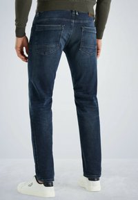 Jean en denim bleu foncé, ajusté, avec deux poches arrière et des détails de couture visibles. Portés avec des baskets blanches.