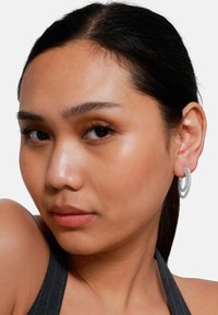 Orecchini a cerchio d'argento con una texture intrecciata, indossati da un modello con un'espressione neutra, che presenta una pelle liscia e capelli scuri.