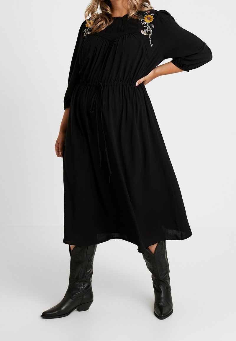 Robe noire midi avec des manches trois-quarts, taille cintrée et détails floraux brodés. Portée avec de grandes bottes de cow-boy noires.