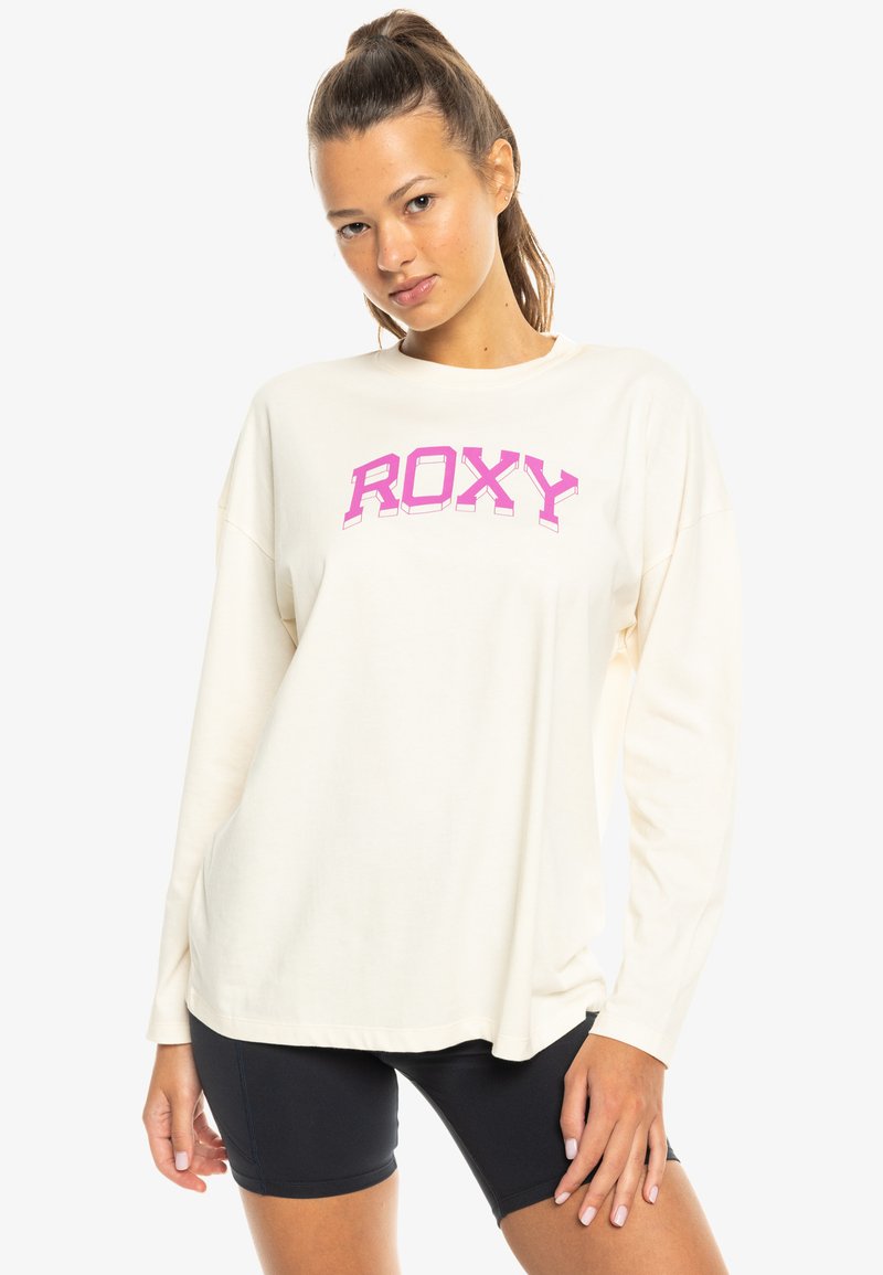 Camisa de manga larga Roxy en blanco roto con un logo rosa llamativo. Corte holgado, tela suave, cuello redondo y hombros caídos. Combinada con pantalones cortos negros.