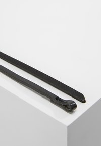 Černé metalové botníky s plochým designem, přičemž jeden má smyčkový konec a druhý se zúženým hrotem, vystavené na bílé ploše.