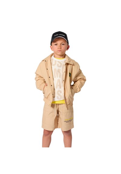 Garçon portant un ensemble veste et short beige, chemise blanche à motifs, et casquette noire Marc Jacobs, debout les mains dans les poches.
