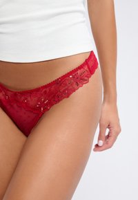 Tanga de encaje rojo con malla transparente y patrón de lunares, con un borde de encaje con forma de onda adornado con lentejuelas y detalles florales.