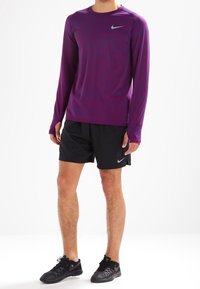 Fioletowa, długozasłonowa koszulka sportowa z srebrnym logo Nike, w połączeniu z czarnymi szortami i czarnymi butami sportowymi. Gładki, lekki materiał.