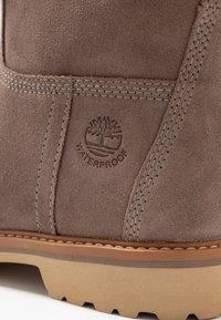 Botte imperméable marron à texture suédée, avec des coutures décoratives et une semelle en caoutchouc. Logo d'arbre en relief et étiquette "IMPERMÉABLE" visibles.