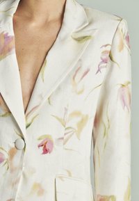 Blazer floral en tela ligera con sutiles patrones en rosa y verde, cierre de un botón, cuello solapa y bolsillos laterales.