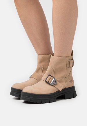 Bottines en suédine beige avec une semelle noire épaisse, présentant une boucle en métal décorative et un branding minimaliste sur le côté.