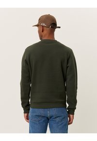 Olivgrüner Pullover mit Rundhalsausschnitt und gerippten Bündchen, kombiniert mit blauen Jeans. Der Stoff wirkt weich mit minimaler Textur.