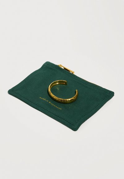 Βραχιόλι μανσέτα με υφή χρυσού που ξεκουράζεται πάνω σε σακούλα από σκούρο πράσινο ύφασμα με χρυσλό φερμουάρ και διακριτικό κείμενο της μάρκας.