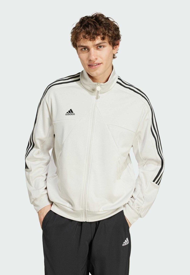 Giacca bianca zip-up con colletto alto, con accenti neri a tre strisce sulle maniche e logo Adidas sul petto. Tessuto morbido.