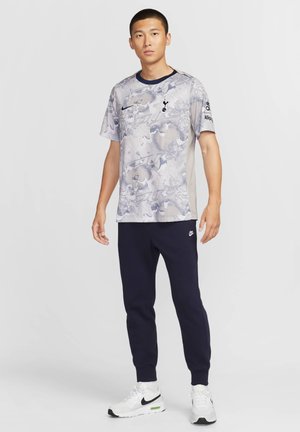 Nike sportkläder som inkluderar en ljusgrå mönstrad kortärmad skjorta med blå accenter och marinblå joggers, tillsammans med vita sneakers.