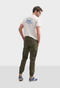 Khaki cargobroeken met zijzakken, gecombineerd met een crèmekleurig t-shirt met blauwe logotekst, en blauwe sneakers met oranje accenten.