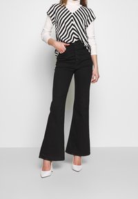 Sorte flared jeans med høj talje og fem-knaps lukning, parret med en sort og hvid stribet, løs pasform top. Skoene er hvide.