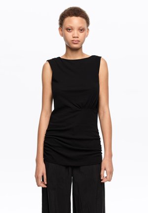 Joven con cabello corto y rizado, vestido con una blusa negra sin mangas y pantalones negros, posando frente a un fondo blanco liso.