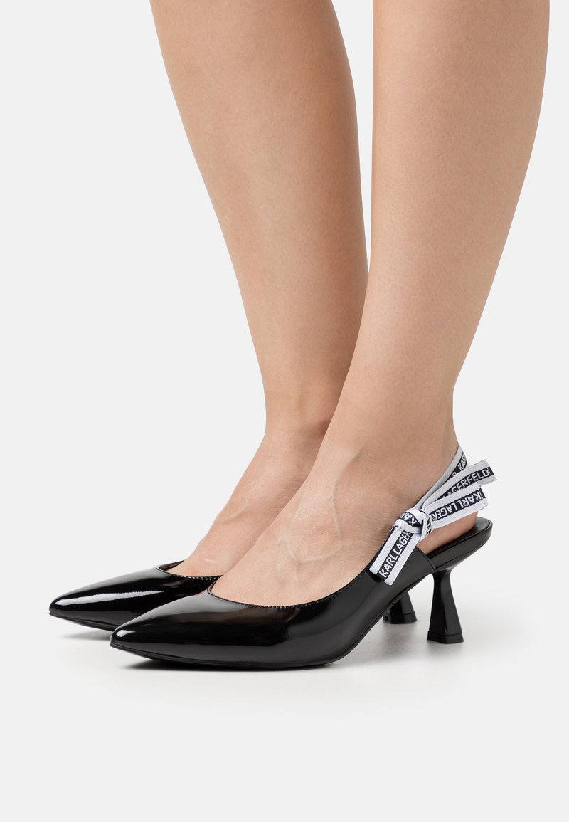 Décolleté in vernice nera con punta affusolata, caratterizzate da un tacco a blocco più corto e una fascia decorativa con nastro e branding.