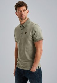 Olivevihreä polo-paita, jossa on teksturoitu kudos, kolme nappia napitus, ja lyhyet hihat. Rinnassa on pieni musta brodeerattu logo.