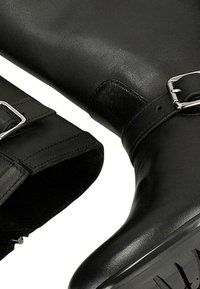 Černé kožené boty s kovovými přezkami a drsnou gumovou podešví, zobrazené zblízka na bílém pozadí.