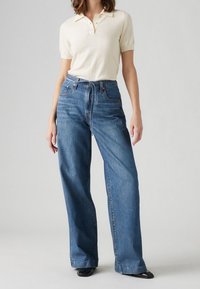 Femme portant un polo crème à manches courtes rentré dans un jean bleu taille haute à jambes larges avec des chaussures noires.