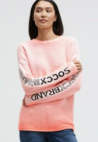 Pink knitted sweater with striped texture and long sleeves featuring "SOCKBRAND" logo in black on the arms. Round neckline.