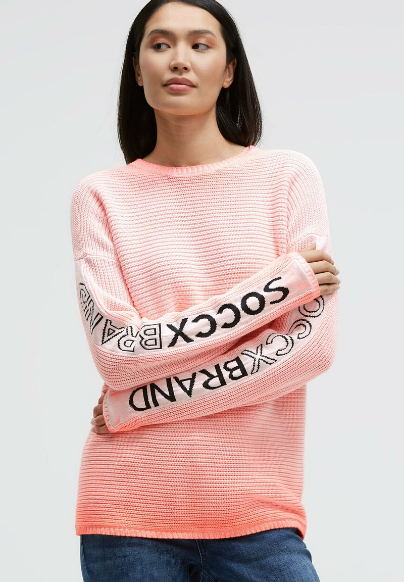 Pink knitted sweater with striped texture and long sleeves featuring "SOCKBRAND" logo in black on the arms. Round neckline.