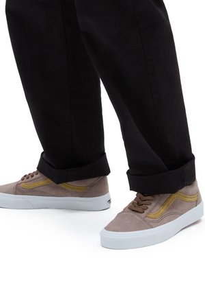 Braune Wildleder-Sneaker mit gelben Akzenten, weißen Gummisohlen und braunen Schnürsenkeln. Kombiniert mit schwarzen, hochgekrempelten Hosen.