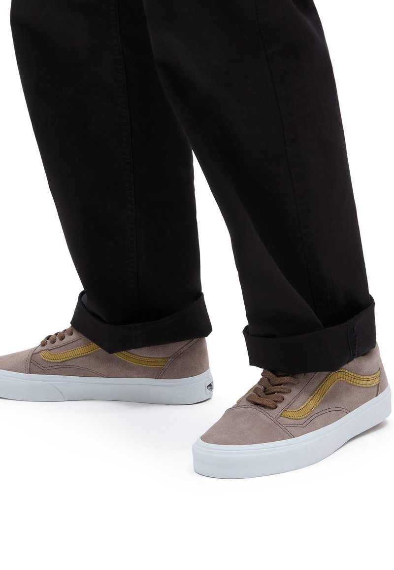 Sneaker in suede marrone con dettagli gialli, suole in gomma bianca e lacci marroni. Abbinati a pantaloni neri arrotolati.