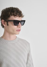 Zwarte geometrische zonnebril met scherpe hoeken en donkere lenzen, gedragen door een persoon in een lichtgrijze gestructureerde trui met een zigzagpatroon.